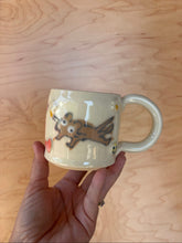 Load image into Gallery viewer, Hand holding a ceramic mug with a cartoon design on a wooden surface

