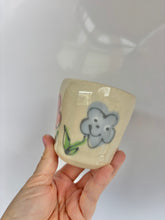 Load image into Gallery viewer, Hand holding a small ceramic cup with a smiley flower design on a plain background

